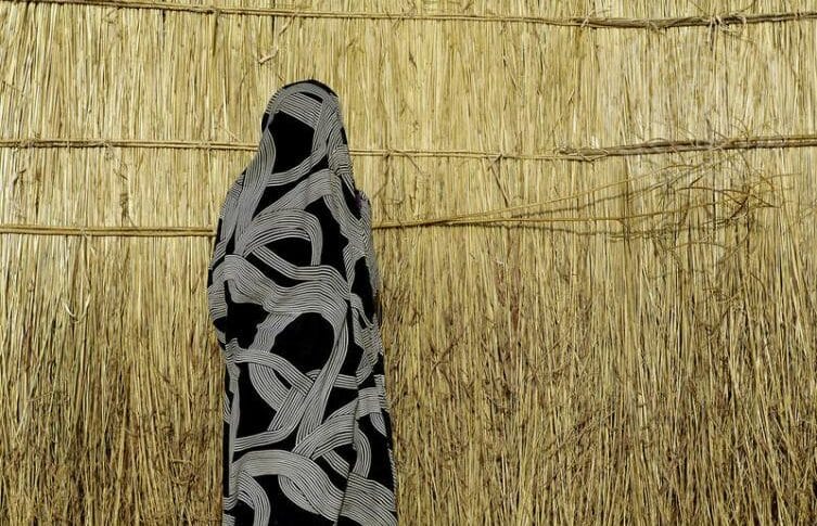 A Misseriya woman stands in front of a thatched shelter
