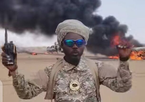 A Joint Force fighter holds a radio in front of burning vehicles following clashes in Sodari, North Kordofan, September 17, 2026.